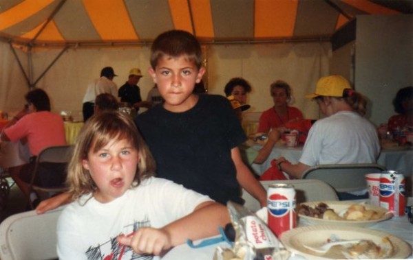 Dominick at age 10 with his best friend, Ron, age 9. Dom, a white presenting person, is wearing a white racing t-shirt with red lettering and has shoulder length, dirty blond hair, and Ron, a very tan Italian, is wearing a black t-shirt with short dark hair. They are in a tent, sitting at a table with food plates and cans of Pepsi on them as they have just eaten a meal at Michigan International Speedway. Dom is licking the corner of his lip when the camera caught him.