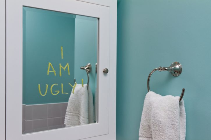 A mirror on the wall reflects a pale toothpaste minty green blue wall with a ring on it, holding a white towel. Written on the mirror are the words I Am Ugly in yellow lettering.