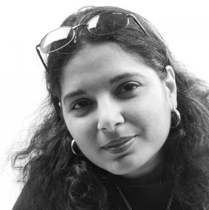 Day, a female identified individual smiles slightly at the camera, long dakr hair around her shoulders, a pair of sun glasses on her head, and she is dressed in black.