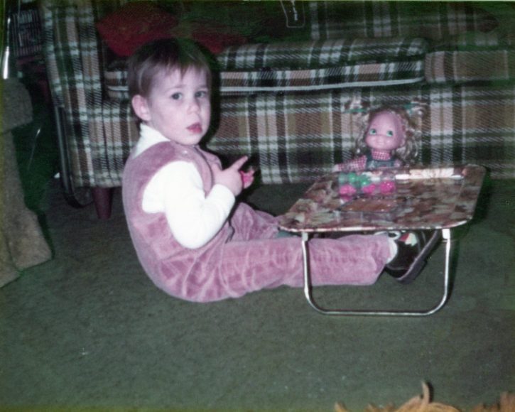 A photo of a toddler with SMA from the early 1980s sitting with a little tray with books on it in front of them, next to a retro plaid sofa, with the doll next to them. They are wearing suede overalls.