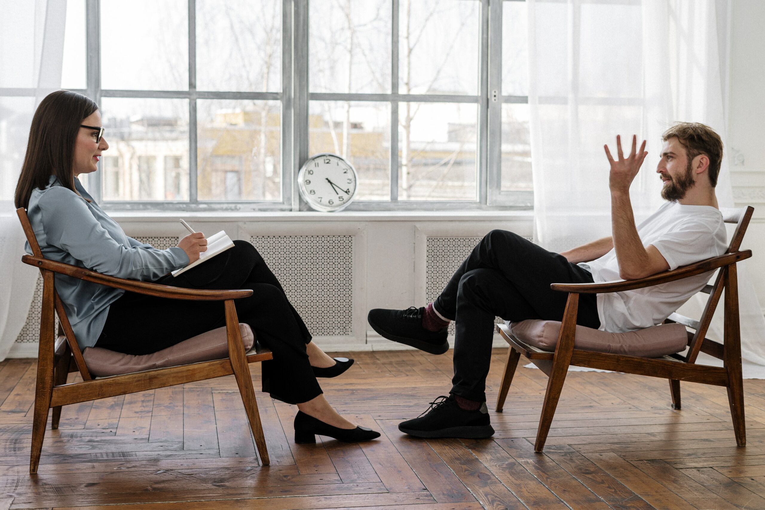 ID: two people sit in a room. There are windows behind them and a clock that reads 5:22 PM. The floor is wooden. They are both sitting in wooden chairs and wearing black pants and black shoes. One is a man leaning back in his chair and he has a beard. The other is a woman. Both appear to be white. The woman is sitting and writing things down in a notebook. She has classes and a blue long sleeve shirt and has long brown hair. He is wearing what looks like a white T-shirt and has dirty blonde/light brown hair.