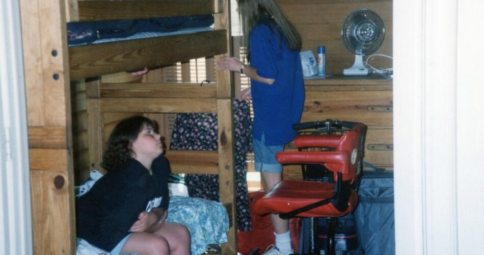A femme presenting disabled person with medium light brown hair sits on a bunk bed. On the lower bunk. They are wearing shorts and a T-shirt. There is another person behind them with their back to the camera.